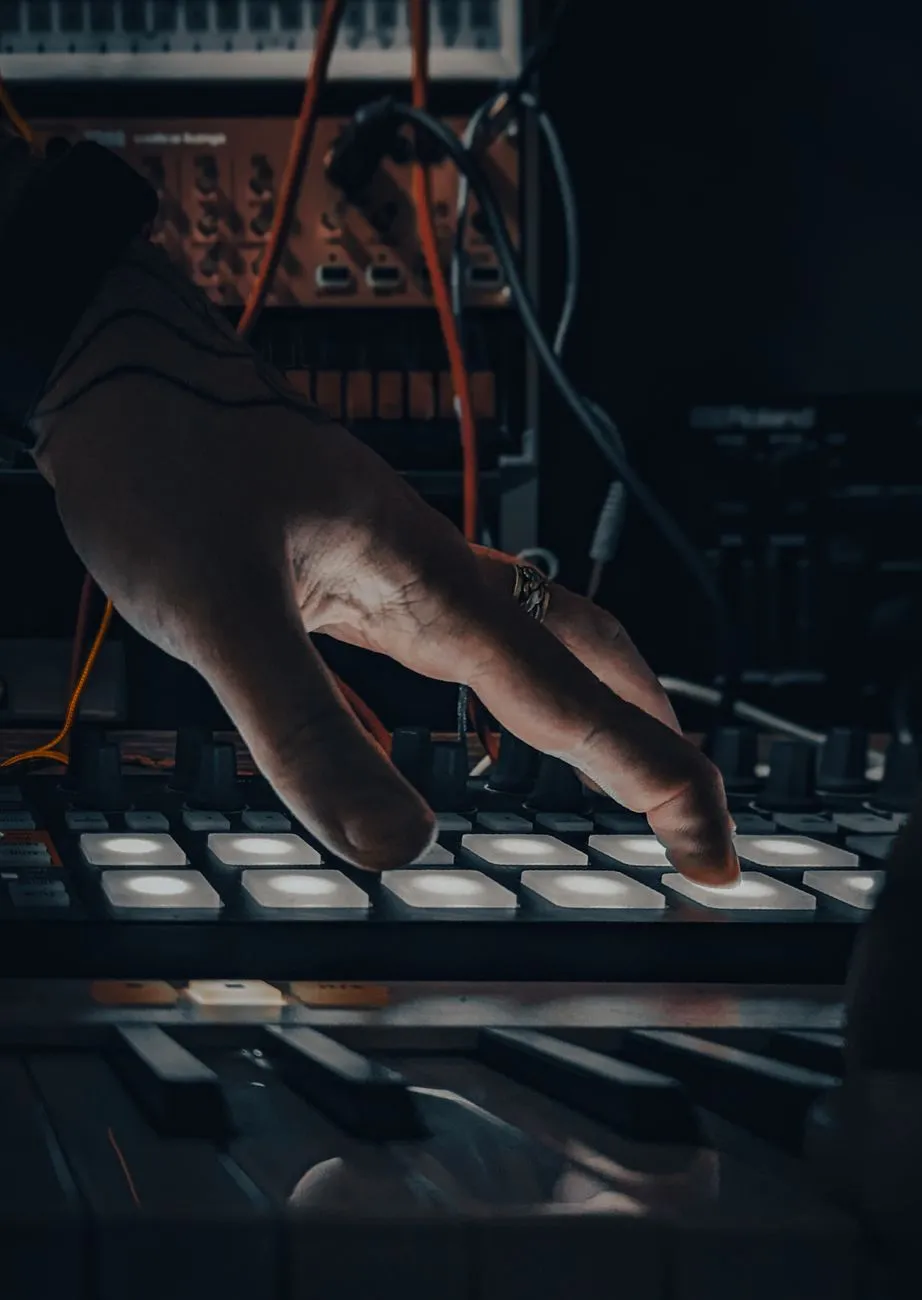 A musician's hand operating a synthesizer pad with glowing buttons in a dark stu